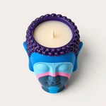 Vela artesanal THE IBIZA CANDLES con forma de cabeza de Buda pintada a mano en tonos azul, violeta y rosa.