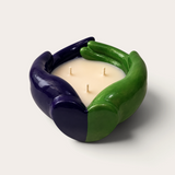 Vela artesanal THE IBIZA CANDLES con forma de manos entrelazadas en colores verde y morado, diseño escultórico.